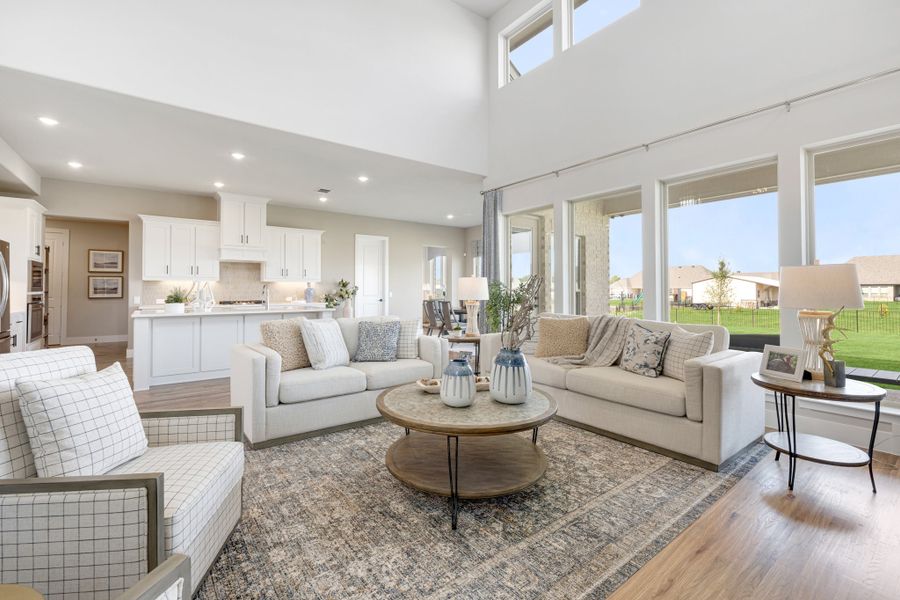 Open-concept living room with high ceilings, large windows, hardwood floors, and white kitchen visible in background.