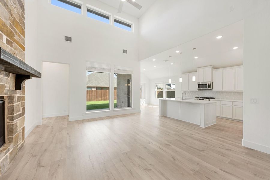 Open-concept living area with stone fireplace, high ceilings, and kitchen with white cabinets and island visible in background