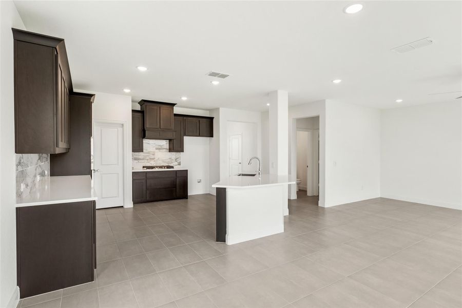 Open kitchen with dark wood cabinets, white island with sink, stone backsplash, and gray tile floors Open kitchen with dark wood cabinets, white island with sink, stone backsplash, and gray tile floors