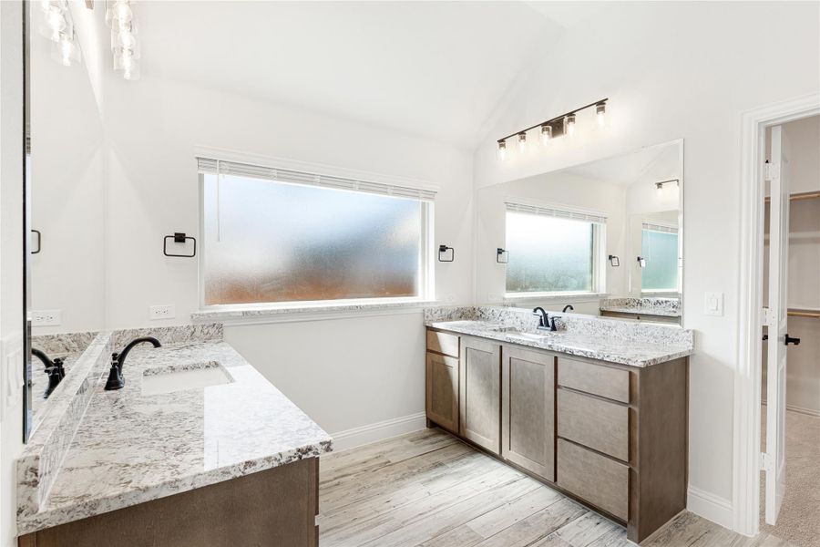 Master bathroom with dual vanities, granite countertops, wood-look floors, and vaulted ceiling with frosted window