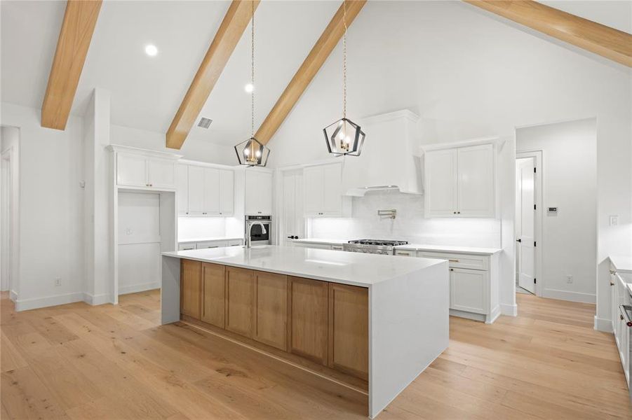 Vaulted ceiling kitchen featuring exposed wood beams, a spacious central island with a waterfall countertop, white shaker cabinetry, light wood-finish flooring, and two modern pendant lights