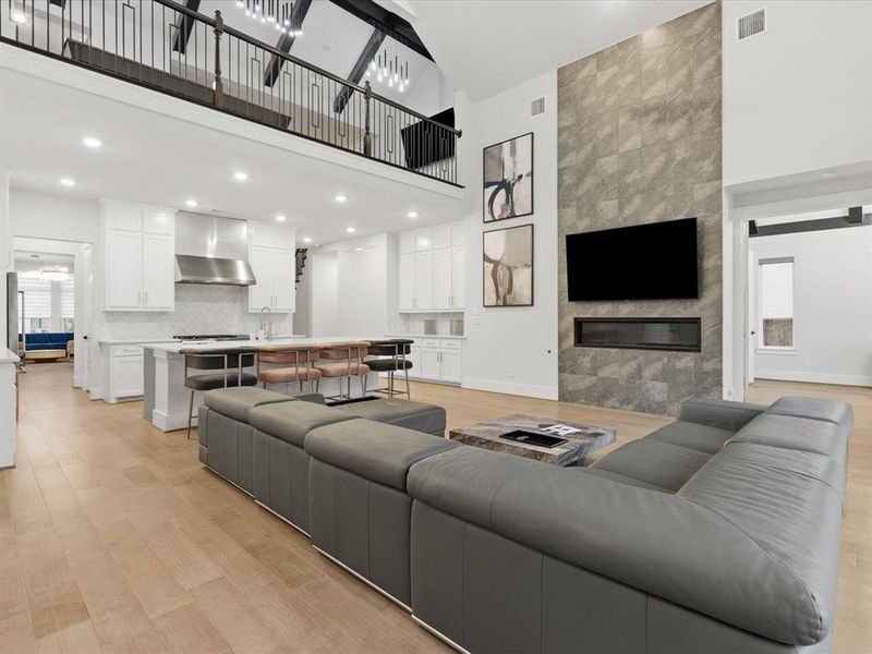 Open-concept living space featuring wood-finish flooring, a two-story tiled fireplace wall with an integrated linear firebox, and a gourmet kitchen with a central island Open-concept living space featuring wood-finish flooring, a two-story tiled fireplace wall with an integrated linear firebox, and a gourmet kitchen with a central island