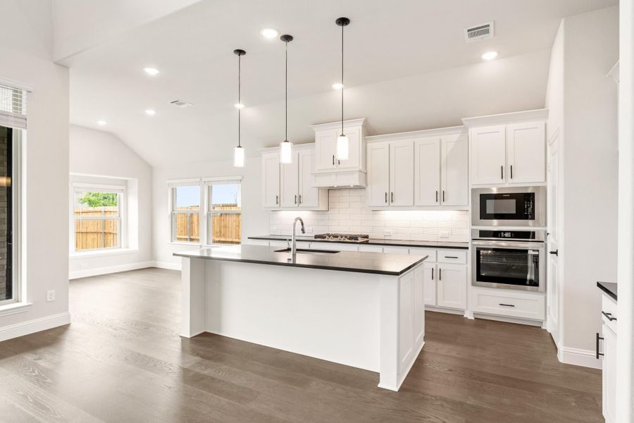 Open kitchen with white cabinets, dark island, stainless steel appliances, and hardwood floors under vaulted ceiling Open kitchen with white cabinets, dark island, stainless steel appliances, and hardwood floors under vaulted ceiling