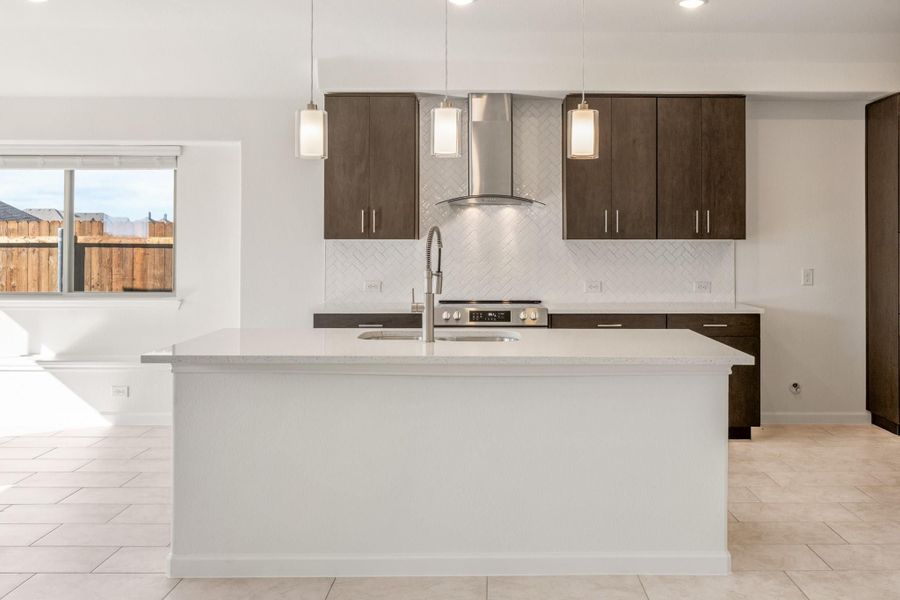 Kitchen with large white island, dark wood cabinets, stainless steel range hood, and pendant lights over herringbone tile backsplash.
