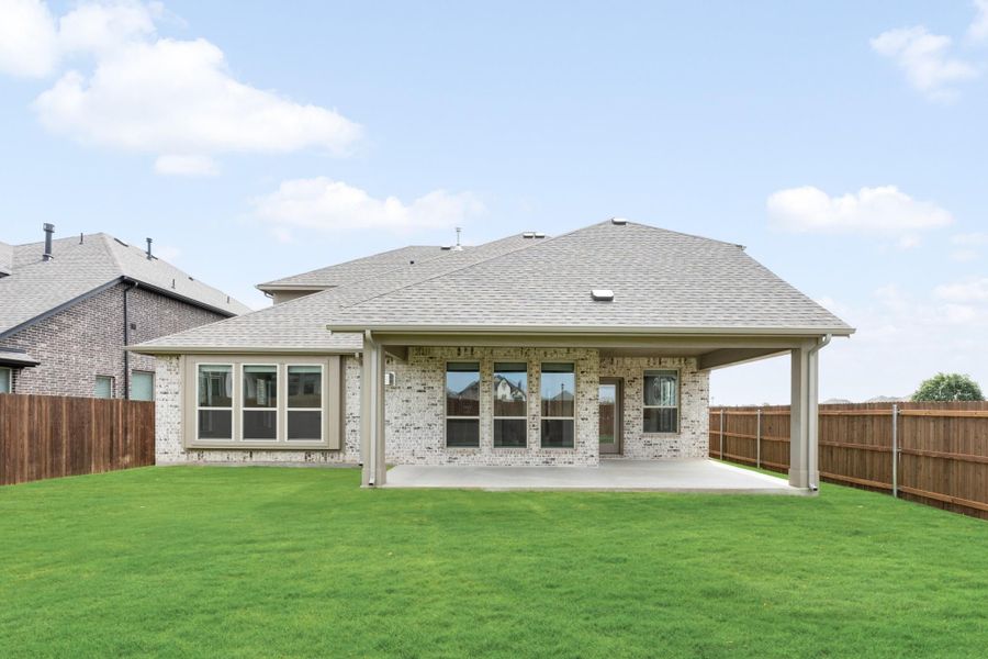 Rear view of brick home with covered patio, large windows, and fenced backyard with green lawn