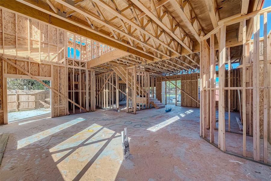 This photo shows a house under construction, featuring wooden framing with visible beams and trusses. The open space suggests a future two-story layout with large windows and ample natural light.