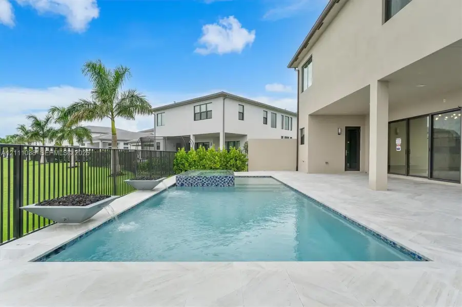 Swimming pool with a raised spa, decorative stone spillover bowls, neutral-toned tiled decking, and a black metal perimeter fence