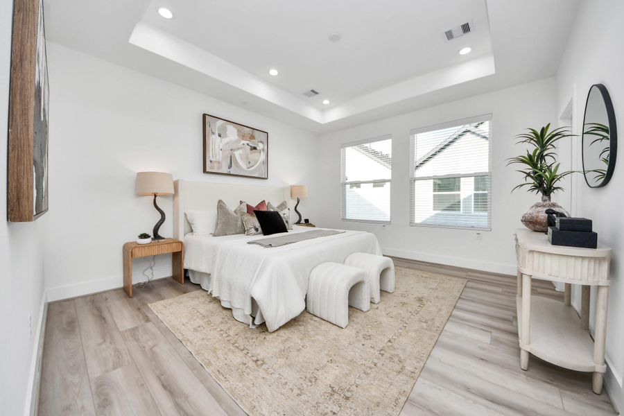 Primary bedroom features a modern, bright design with large windows, a tray ceiling, and stylish recessed lighting. It includes a cozy bed, contemporary furnishings, and neutral decor, creating a serene and inviting atmosphere.