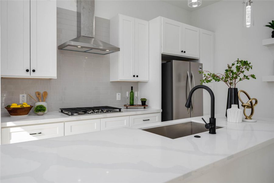 A sleek, modern kitchen features crisp white cabinetry, stainless steel appliances, and a spacious quartz island accented by a matte black faucet and fresh greenery.