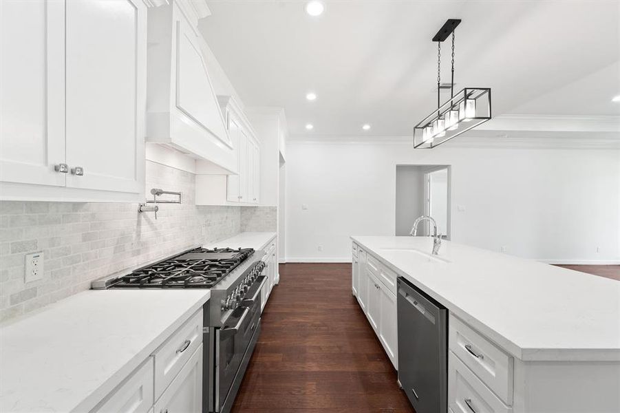 Modern kitchen featuring white cabinetry, stainless steel appliances, gas stove, and a large island with a sink. It has recessed lighting, a stylish pendant light fixture, and dark hardwood floors.