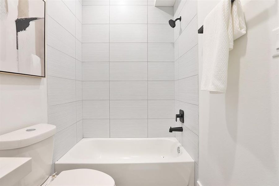 This is a modern bathroom featuring a combination of bathtub and shower with sleek black fixtures. It has white textured wall tiles and includes a toilet with an adjacent mounted towel rail. A framed abstract art piece adds a touch of elegance to the space.