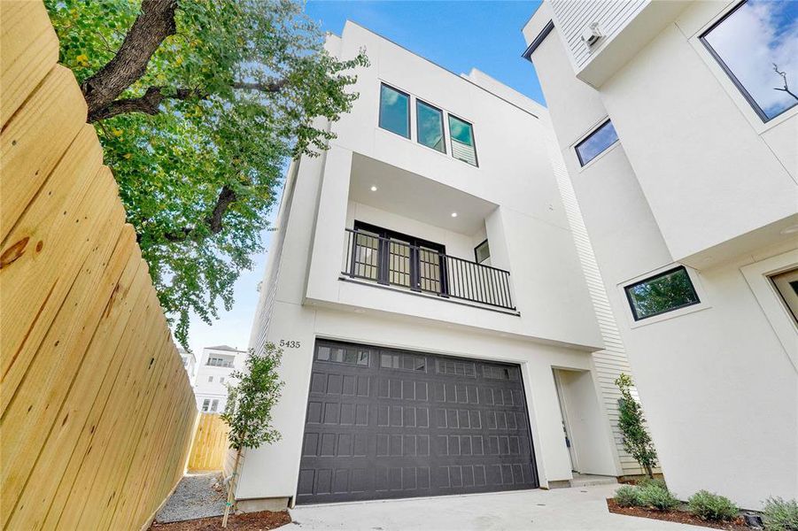 Modern three-story home with a sleek stucco exterior, featuring a large garage door, balcony, and minimalistic landscaping. Located in a cozy neighborhood with a wooden privacy fence and mature trees.