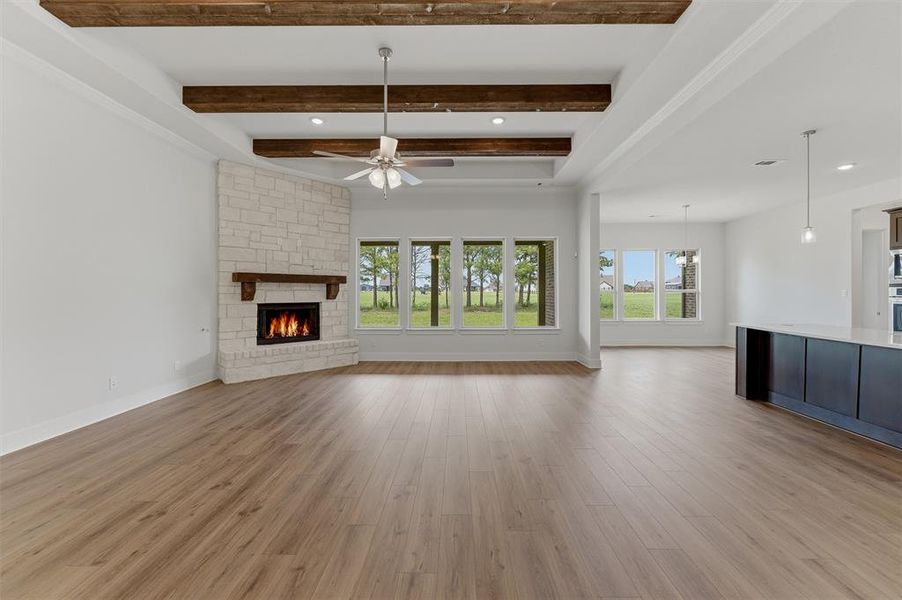 Spacious living area featuring wood-finish flooring, a stone-clad fireplace with a wood mantel, exposed ceiling beams, a ceiling fan, and multiple windows providing ample natural illumination