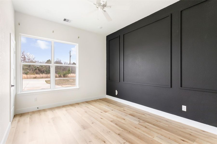 This primary bedroom features large windows for natural light, light wood flooring, and a striking black accent wall with decorative paneling, offering a modern and stylish space.