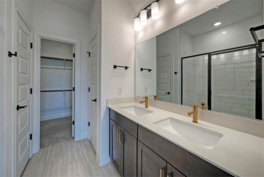 Double vanities with gold fixtures pair beautifully with tile floors in this modern primary bathroom. Double vanities with gold fixtures pair beautifully with tile floors in this modern primary bathroom.