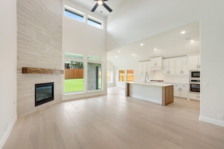 Open-plan living area with tiled fireplace, high ceilings, and kitchen with white cabinets and island visible in background Open-plan living area with tiled fireplace, high ceilings, and kitchen with white cabinets and island visible in background