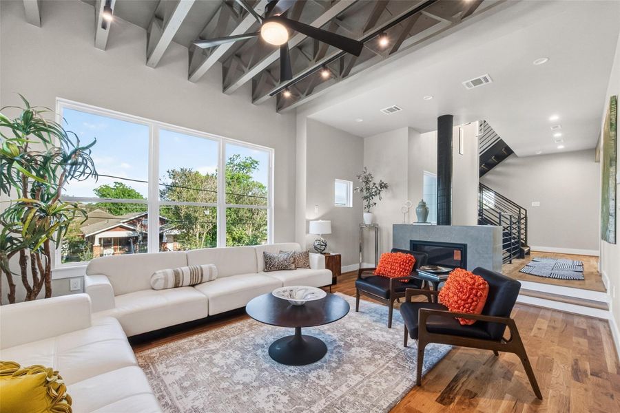 The living room offers generous proportions, hardwood floors, a 42" free-standing gas fireplace, and a wall of windows with tree views. The open-riser staircase adds architectural drama, and the volume created by the exposed truss ceiling makes this space feel expansive and airy.