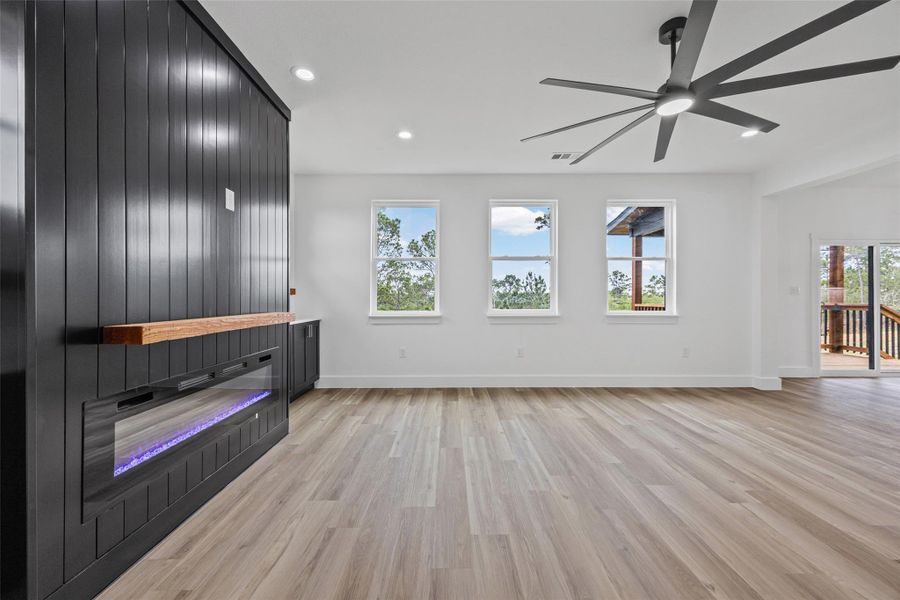 Spacious living area featuring light wood-finish flooring, a modern electric fireplace with a wood mantle, and a contemporary ceiling fan Spacious living area featuring light wood-finish flooring, a modern electric fireplace with a wood mantle, and a contemporary ceiling fan
