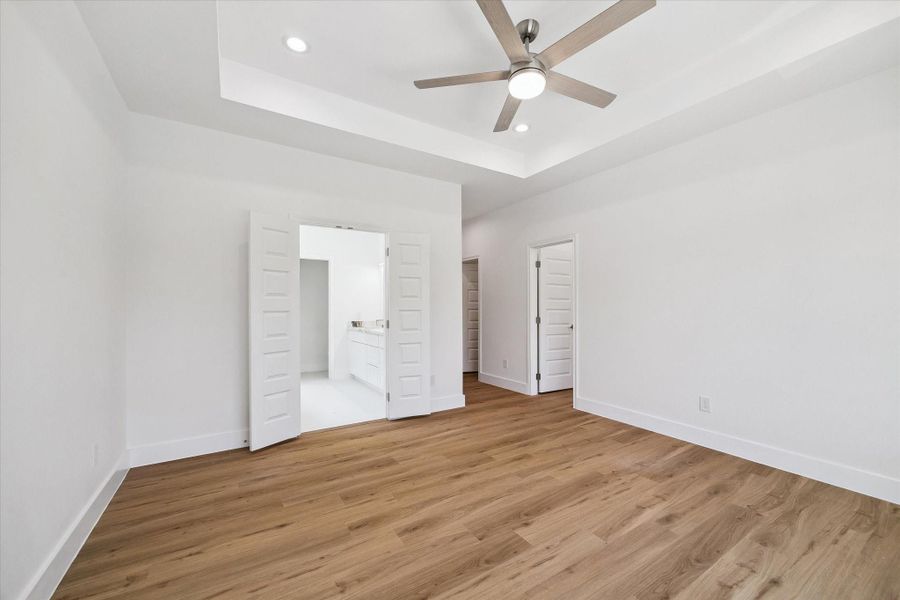 The spacious primary bedroom offers a clean, modern retreat with warm wood-look flooring, crisp white walls, and a dramatic tray ceiling. A contemporary ceiling fan and recessed lighting add comfort and style, while double doors provide direct access to the adjoining bath. Generous proportions allow plenty of room for a complete bedroom suite, seating area, or additional furnishings.
