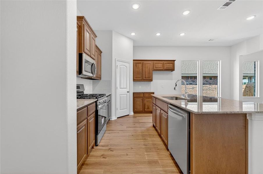 Kitchen with light stone counters, stainless steel appliances, brown cabinetry, a center island with sink, and recessed lighting