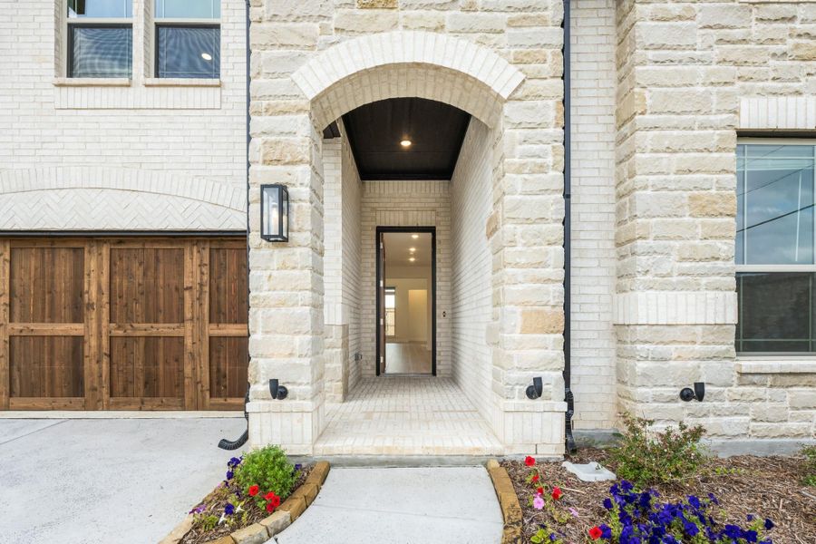Stone and brick home entrance with arched doorway, dark wood garage door, and small flower beds along the walkway