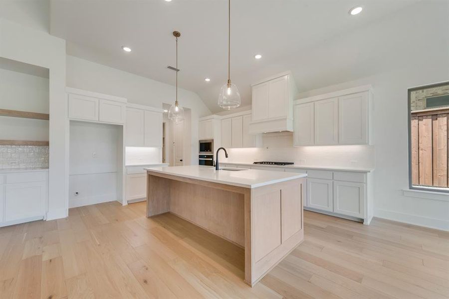 Kitchen with decorative backsplash, decorative light fixtures, light wood-style floors, white cabinetry, and an island with sink