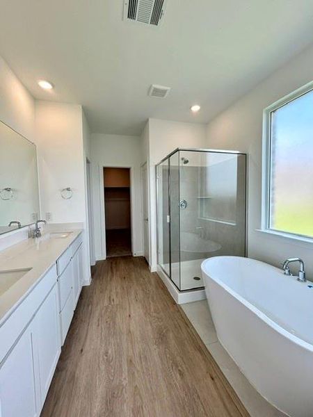 Bathroom featuring a freestanding soaking tub, glass-enclosed shower, dual vanity with white cabinetry and light countertops, and wood-finish flooring Bathroom featuring a freestanding soaking tub, glass-enclosed shower, dual vanity with white cabinetry and light countertops, and wood-finish flooring