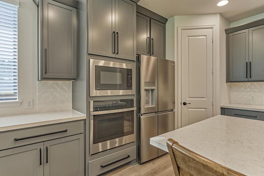 This modern kitchen features sleek gray cabinets, stainless steel appliances, and a spacious refrigerator. The countertop is a light, durable surface, complemented by a stylish herringbone tile backsplash. Natural light streams through a nearby window, enhancing the room's inviting ambiance.
