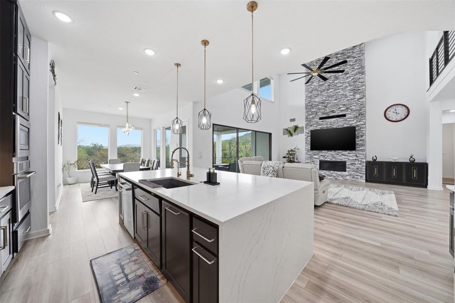 The expansive open-concept living area features light-colored flooring, a kitchen island with a dark sink, and a living room with a stone-accented wall and a ceiling fan