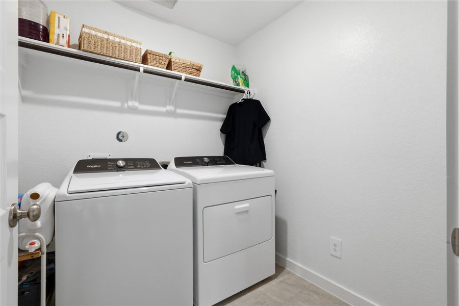 This laundry room features a modern washer and dryer set, ample shelving for storage, and a clean, bright aesthetic with white walls and tile flooring.