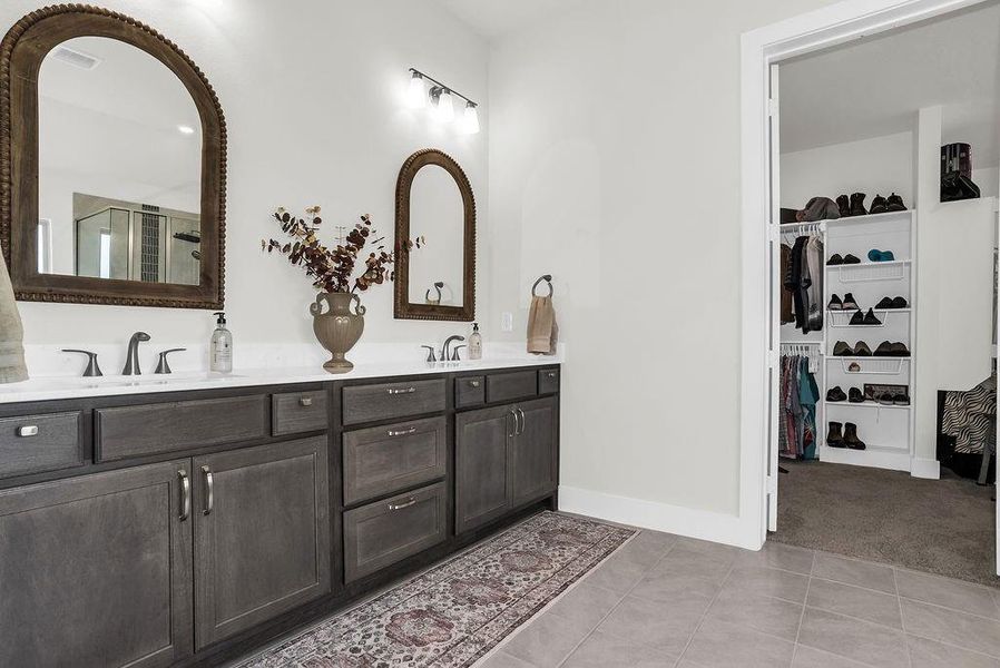 Main ensuite bath features dual vanity with wood-finish cabinetry, brush bronze faucets,  glass-enclosed steam shower with a tiled surround and vertical accent, a freestanding soaking tub ceramic tile floors and spacious walk-in closet with built-in shelving.