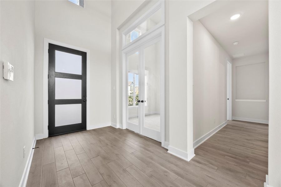 This photo showcases a modern entryway with light wood flooring, a sleek black front door with frosted glass panels, and high ceilings. There's a hallway leading to additional rooms and a set of glass double doors, allowing natural light to brighten the space. This photo showcases a modern entryway with light wood flooring, a sleek black front door with frosted glass panels, and high ceilings. There's a hallway leading to additional rooms and a set of glass double doors, allowing natural light to brighten the space.
