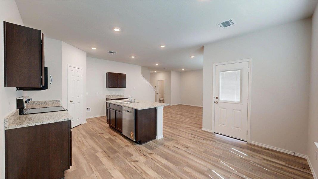 Open-concept kitchen and living area featuring wood-finish flooring, dark wood cabinetry, light granite countertops, a kitchen island with integrated sink and dishwasher, and recessed lighting Open-concept kitchen and living area featuring wood-finish flooring, dark wood cabinetry, light granite countertops, a kitchen island with integrated sink and dishwasher, and recessed lighting