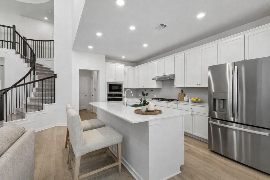 Spacious kitchen anchored by an oversized quartz island with seating, stainless steel appliances, and a gas cooktop with vent hood. Seamless flow to the adjacent living area and striking curved staircase creates a functional layout ideal for everyday living and entertaining.
