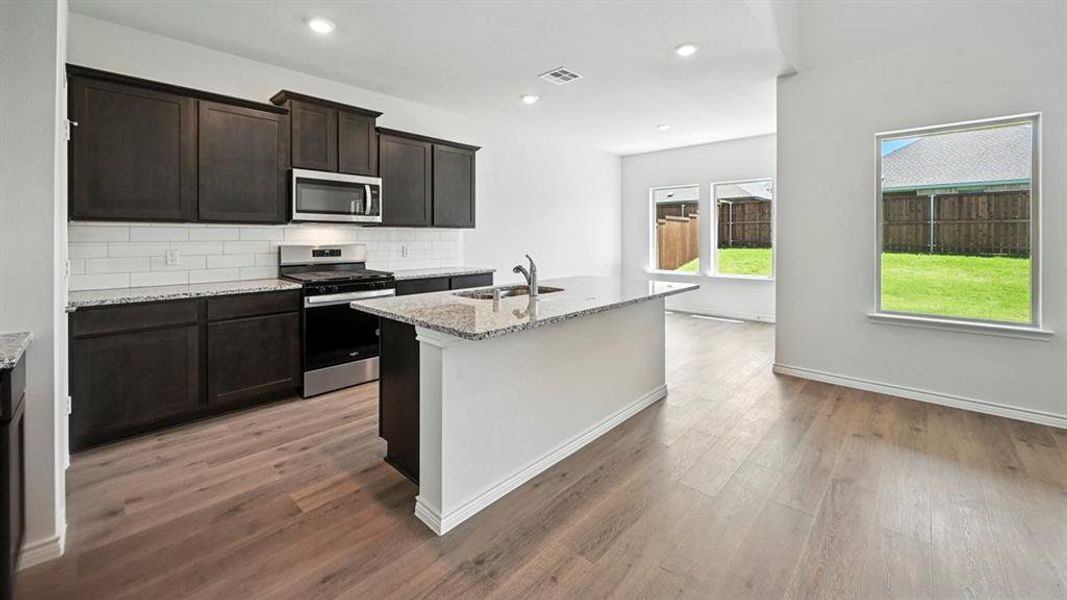 Kitchen featuring stainless steel appliances, light stone countertops, an island with sink, tasteful backsplash, and light wood-type flooring