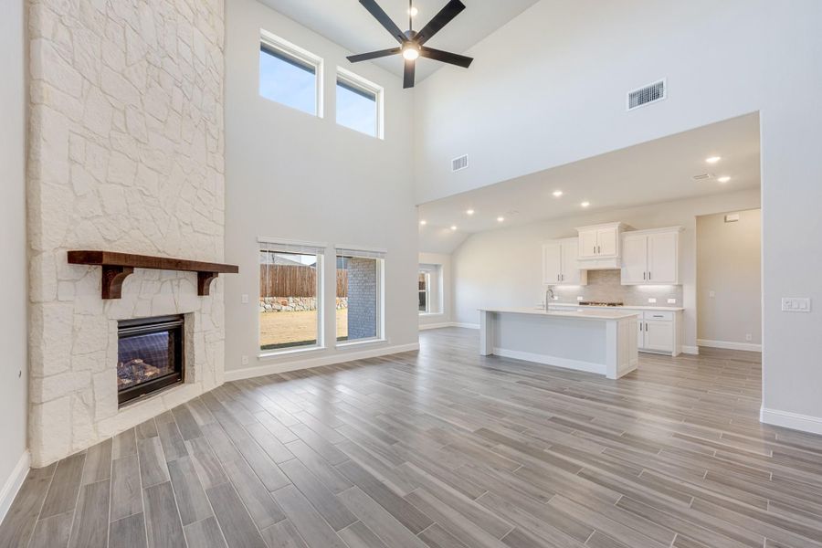Open living area with tall stone fireplace, wood-look floors, and open kitchen with white cabinets in background Open living area with tall stone fireplace, wood-look floors, and open kitchen with white cabinets in background