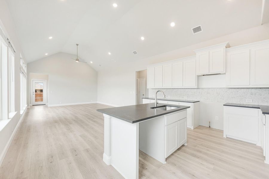 Open kitchen with white cabinets, dark countertops, center island, and light wood floors opening to living area with vaulted ceiling