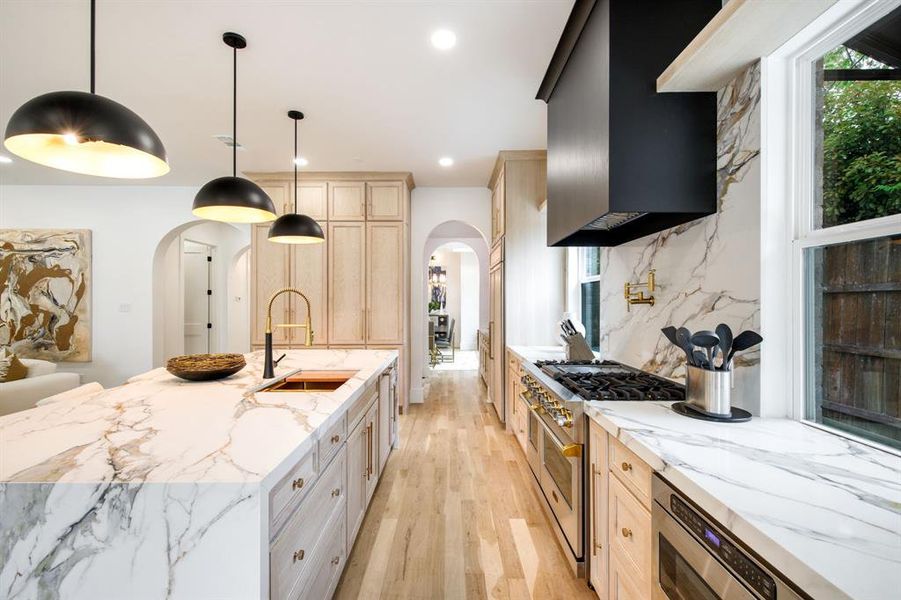 Gourmet kitchen featuring a substantial island with integrated sink, luxurious stone countertops, light wood-finish cabinetry, a professional-grade range, and contemporary pendant lighting Gourmet kitchen featuring a substantial island with integrated sink, luxurious stone countertops, light wood-finish cabinetry, a professional-grade range, and contemporary pendant lighting