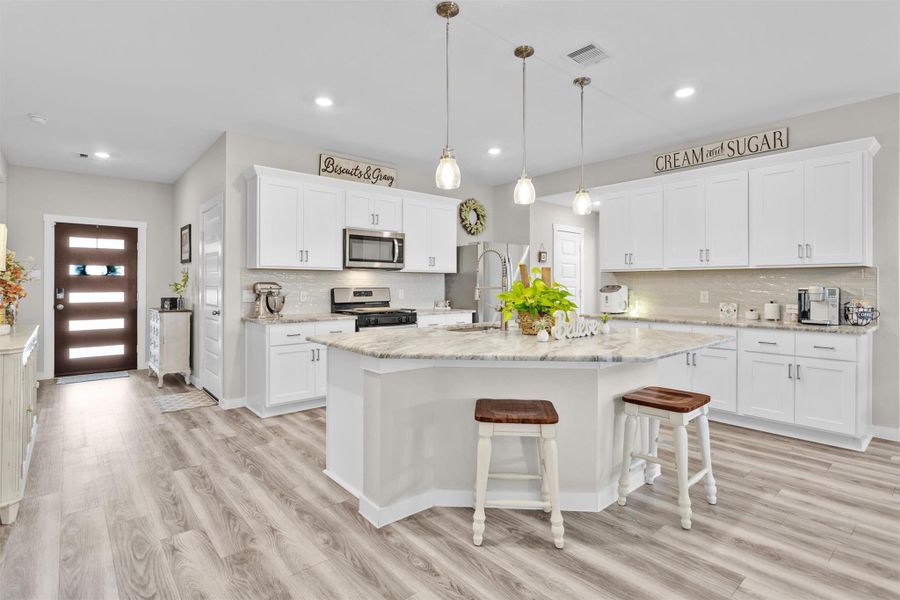 This modern kitchen features white cabinetry, stainless steel appliances, and a spacious island with leathered granite countertops. The open layout is complemented by wood-style flooring and pendant lighting, creating a bright and inviting space.