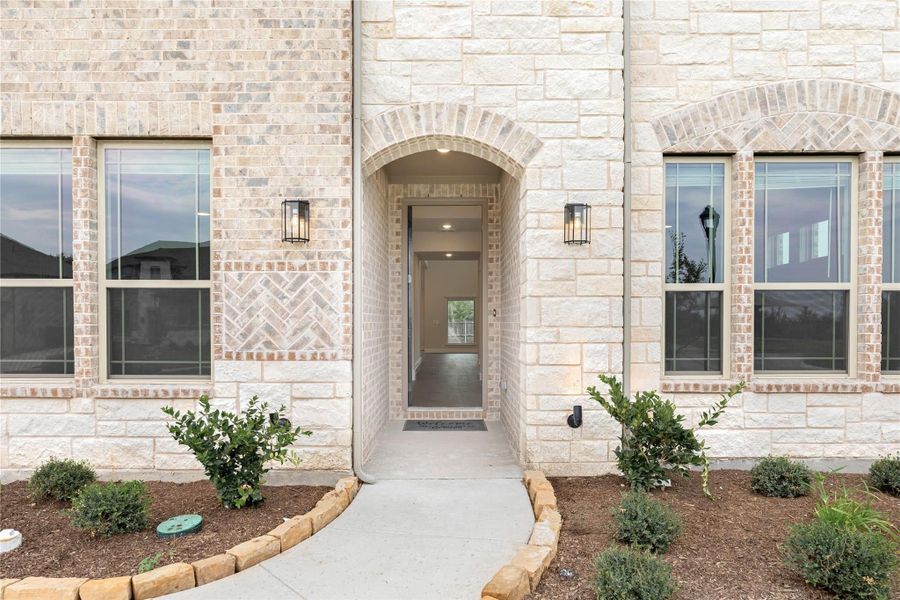 Stone and brick home entrance with arched doorway, wall sconces, and landscaped front walkway Stone and brick home entrance with arched doorway, wall sconces, and landscaped front walkway