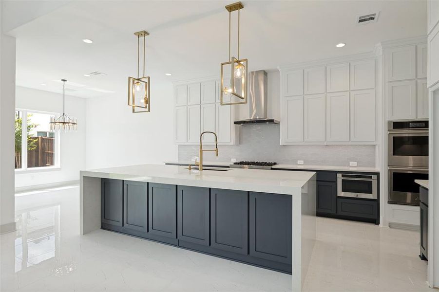 Open-concept kitchen featuring a large island with dark cabinetry, white countertops, stainless steel appliances, and two brass pendant lights Open-concept kitchen featuring a large island with dark cabinetry, white countertops, stainless steel appliances, and two brass pendant lights