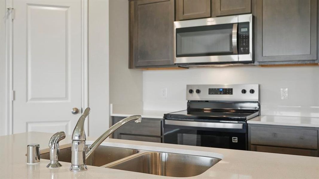 Kitchen featuring a double-basin stainless steel sink with a chrome faucet, light-toned countertops, dark wood-finish cabinetry, a built-in stainless steel microwave, and a stainless steel range