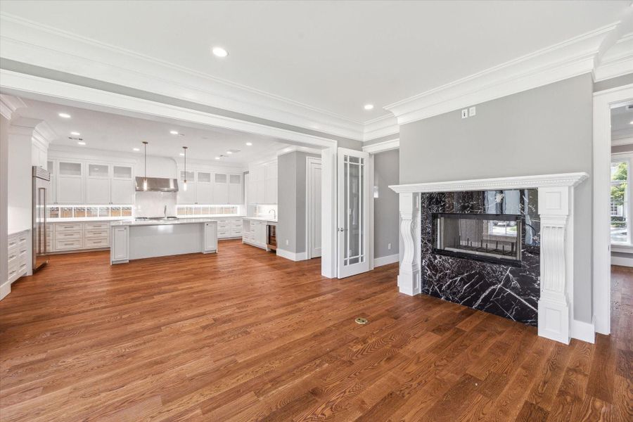 This photo showcases the spacious open-plan kitchen and living area with elegant hardwood floors, a double sided fireplace, with black marble surround and custom mantle. The kitchen features a large island and stainless steel appliances, creating a bright and inviting space ideal for entertaining.