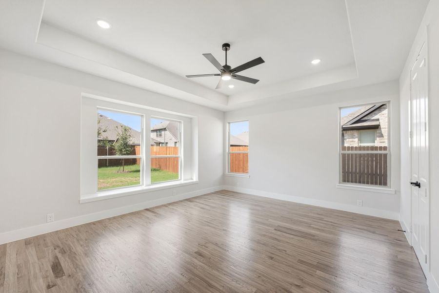 Empty bedroom with tray ceiling, ceiling fan, hardwood floors, and multiple windows with backyard views