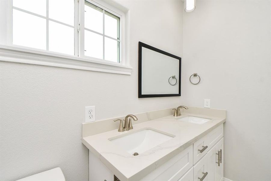 This is a bright, clean bathroom featuring a double sink vanity with modern fixtures and a large mirror, set against a neutral color palette. Natural light streams in from a window above the vanity.