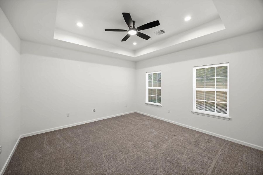 Spacious room featuring a tray ceiling, modern ceiling fan, and plush carpeting, illuminated by recessed lighting and natural light from two large windows.