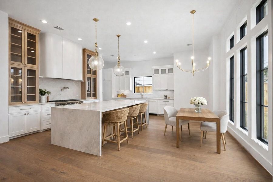 Chef inspired kitchen anchored by a waterfall quartz island, custom white cabinetry, and warm wood accents that add depth and contrast. Glass front upper cabinets, brass pendant lighting, and wide plank hardwood floors create a bright open concept layout with seamless flow to the dining area and expansive black framed windows. *Virtually Staged*