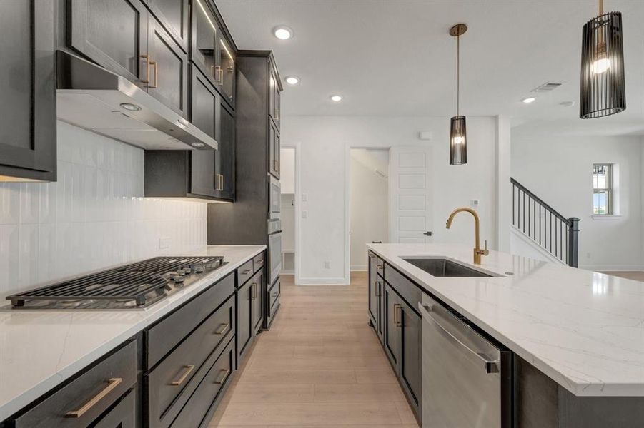 Modern kitchen featuring a gas cooktop, stainless steel hood, quartz countertops, a large island with an undermount sink, and dark cabinetry with brass hardware Modern kitchen featuring a gas cooktop, stainless steel hood, quartz countertops, a large island with an undermount sink, and dark cabinetry with brass hardware