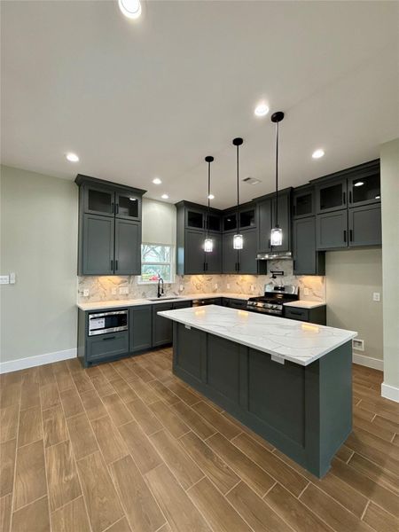This modern kitchen features dark cabinetry, a central island with a Quartz countertop, and stylish pendant lighting. Equipped with stainless steel appliances, it has a sleek backsplash and wood-look tile flooring, offering a blend of elegance and functionality.
