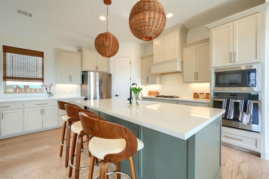 Kitchen with light wood-type flooring, an island with sink, hanging light fixtures, stainless steel appliances, and decorative backsplash